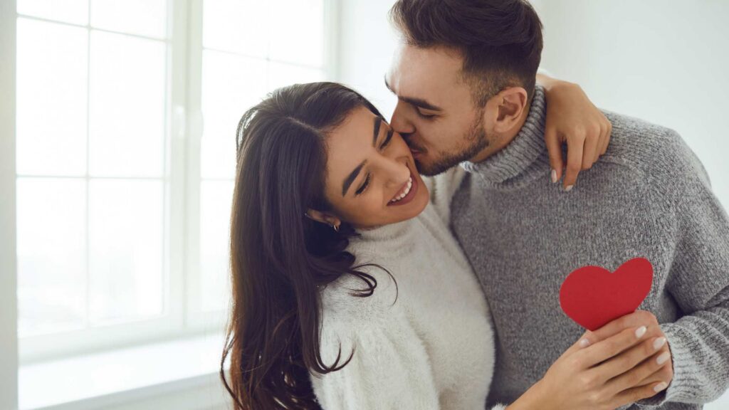 A smiling couple sharing a warm hug in a bright room, holding a small red heart to celebrate a sober Valentine´s Day
