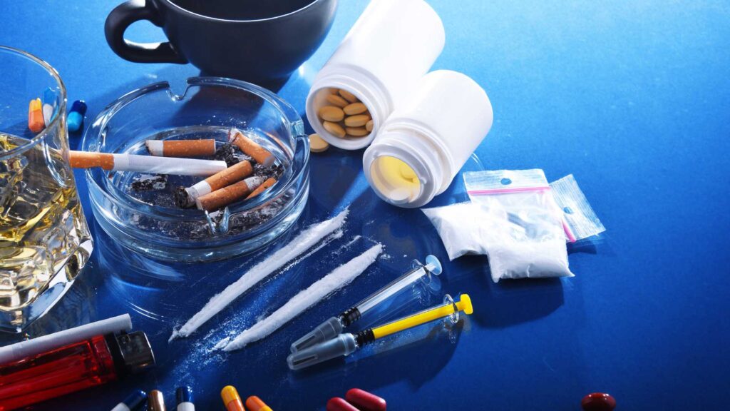 Substance use disorder (SUD): A table with alcohol, cigarettes, pills, bottles, baggies, lines of a white powder, and needles.