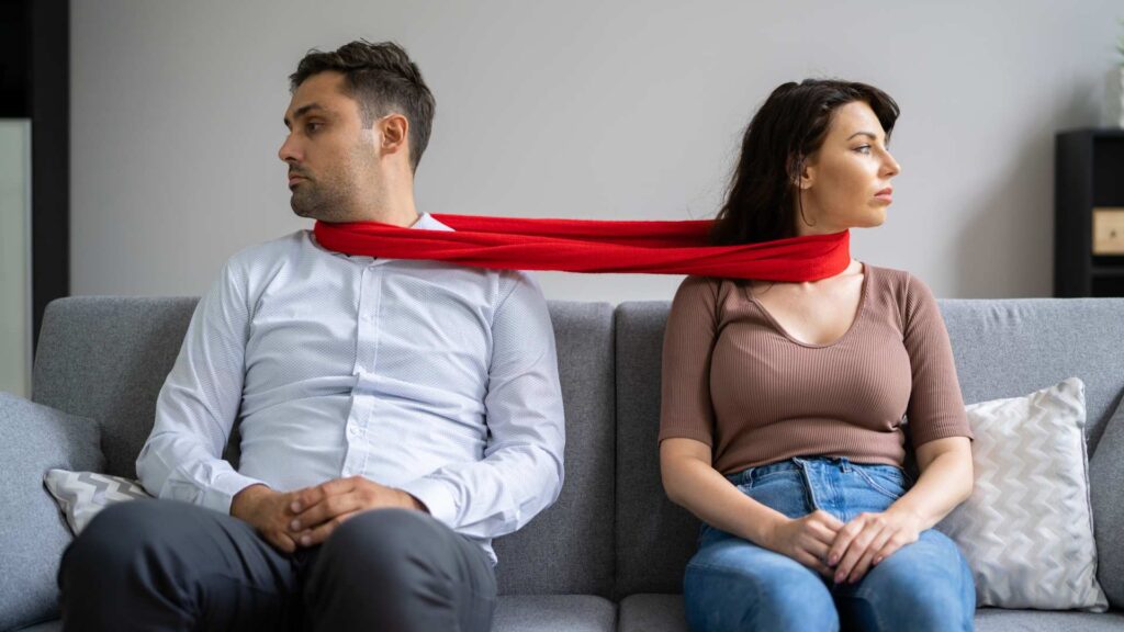 Couple facing away from each other while tied together by a red scarf, hinting at signs of codependency.